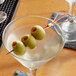 A martini glass with a red bamboo heart skewer holding green olives.