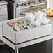 A stainless steel flatware organizer with gray perforated plastic cylinders holding a variety of condiments.