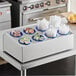 A white table with a white box containing a blue flatware organizer with white and orange yogurt in blue bowls.