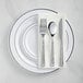 A white plastic plate with banded silver plastic dinnerware and hammered flatware on it.