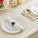 A white table set with Visions gold banded plastic dinnerware and rolled hammered flatware.
