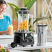 A commercial blender set with a black base, a clear plastic jar filled with fruit, a stainless steel jar, and a tamper tool.