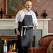 A man wearing a black Choice bib apron with natural webbing accents and holding a glass of wine.