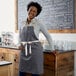 A woman wearing a gray Choice poly-cotton apron with natural webbing accents stands in front of a counter.