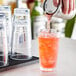 A hand pouring orange liquid from a green bottle into a Acopa Tritan plastic glass filled with ice.