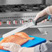 A hand holding a Schraf serrated slicing knife with a blue TPRgrip handle cutting salmon on a blue cutting board.