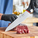 A person wearing black gloves uses a Schraf chef knife to cut a piece of meat on a wooden surface.