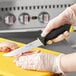 A person in gloves using a Schraf utility knife with a yellow and black TPRgrip handle to cut raw meat on a cutting board.