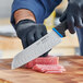 A person in gloves uses a Schraf Santoku knife with a blue TPRgrip handle to cut meat on a cutting board.