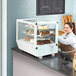 A woman standing behind a white Avantco countertop bakery display case filled with cakes and cupcakes.