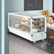 A woman standing behind a white Avantco countertop bakery display case filled with cakes.