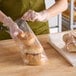 A person in a green apron using a Choice bread bag wicket dispenser to put a bagel in a plastic bag.