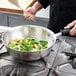 A person cooking vegetables in a Vollrath stainless steel saucier pan with a black and white handle.