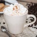 A Tuxton Europa eggshell cappuccino mug filled with coffee and whipped cream with a spoon next to it.
