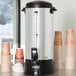 A Proctor Silex coffee urn on a counter with several cups.