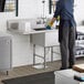 A woman in a kitchen wearing gloves and an apron washing dishes in a Regency stainless steel commercial sink with a drainboard.