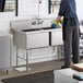 A person in a blue apron washing a Regency stainless steel two compartment sink.