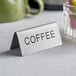 A double sided stainless steel table tent sign that says "Coffee" on a table next to a coffee cup.