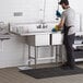 A man in a professional kitchen cleaning a Regency stainless steel 2 compartment sink with a left drainboard.