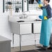 A woman in a blue apron washing dishes in a Regency two compartment sink.