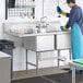 A man in a blue uniform and gloves working in a Regency stainless steel 2 compartment sink with a drainboard.