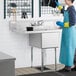 A woman in a blue apron using a Regency stainless steel one compartment sink with drainboards.
