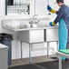 A man in a blue uniform and gloves washing dishes in a school kitchen using a Regency two compartment sink.
