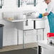A man in a blue uniform washing dishes in a Regency stainless steel 2 compartment sink with a drainboard.