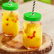 Two Acopa Rustic Charm drinking jars filled with yellow liquid and straws with a green metal lid on a table.