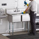 A man in a black shirt and blue jeans using a Regency 2 compartment stainless steel sink in a kitchen.