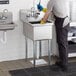 A man in a blue shirt and apron cleaning a Regency stainless steel commercial sink with yellow gloves.