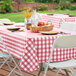 A picnic table with a red and white checkered vinyl table cover, white plates, and a basket of fruit.