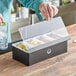 A person using an American Metalcraft matte black condiment bar to hold containers of food.