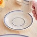 A hand holding an Acopa ivory stoneware cup over a saucer with blue trim.