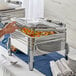 A hand using a silver spoon to serve food from a silver Acopa Manchester chafer.