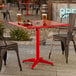A red square aluminum outdoor dining table with an umbrella hole, shown with two metal chairs.