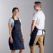 A man and woman wearing navy blue bib aprons with black accents.