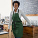 A woman standing in a Choice hunter green adjustable bib apron with black webbing accents.