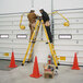 A man in brown overalls and a tool belt working on a yellow Bauer 2-way step ladder with another man.