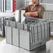 A man in a chef's uniform pouring metal chafing dishes into a grey Choice stackable tote with an attached lid.