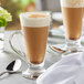 A glass cup of Capora Spiced Chai Latte with a brown liquid in it on a table with a spoon