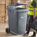 A woman in a yellow reflective vest stepping on a grey Lavex step-on attachment for a wheeled trash can.