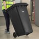 A person in a yellow vest pushing a large black Lavex rectangular trash can.