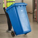 A person in a yellow vest pushing a Lavex blue wheeled rectangular recycle bin.