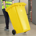 A woman in a yellow shirt pushing a Lavex yellow wheeled rectangular trash can.