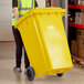 A woman in a yellow vest pushing a Lavex yellow wheeled rectangular trash can.