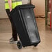 A person in a yellow vest pushing a Lavex black wheeled rectangular trash can.