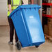 A woman in a yellow vest pushing a Lavex blue wheeled rectangular trash can.