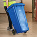 A man in a yellow vest pushing a Lavex blue wheeled rectangular recycle bin.