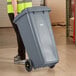 A person in a yellow vest pushes a large grey Lavex rectangular trash can with wheels.